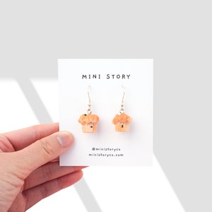 Puede incluir: Un par de pendientes de aro de plata con un amuleto de magdalena en miniatura. Las magdalenas son de color marrón claro con chispas de chocolate marrón oscuro. Los pendientes se muestran en una tarjeta blanca con el texto "MINI STORY" y el nombre de usuario de las redes sociales "@ministoryco" y el sitio web "ministoryco.com".
