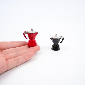 May include: Miniature red and black espresso makers with silver accents. The red espresso maker is held in a hand. The espresso makers have a classic design with a handle and a lid. The background is white.