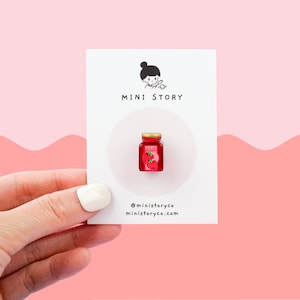 May include: A small, red glass jar with a white lid and the word "JAM" printed on the lid. The jar is filled with red jam and has a strawberry on the front. The jar is on a white card with the words "MINI STORY" printed on it.