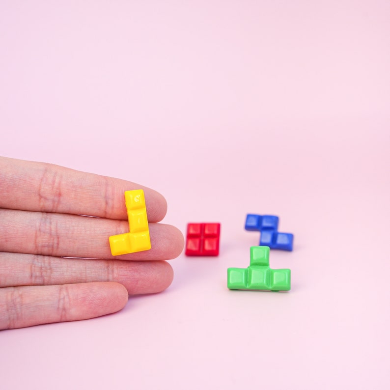 May include: A hand holds a yellow Tetris-shaped block. Three other blocks are on a pink surface: a red square, a blue L-shape, and a green T-shape.