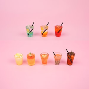 Peut inclure: Un ensemble de huit verres miniatures en verre remplis de liquides de différentes couleurs, notamment vert, orange, rouge et marron. Les verres sont disposés en deux rangées de quatre, avec une paille noire dans chaque verre. Les verres sont sur un fond rose.