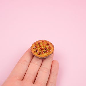 Peut inclure: Une petite tarte avec une croûte en treillis, dans une petite boîte en métal cannelée. La tarte a une croûte brun doré et une garniture verte. La tarte est tenue sur un fond rose.