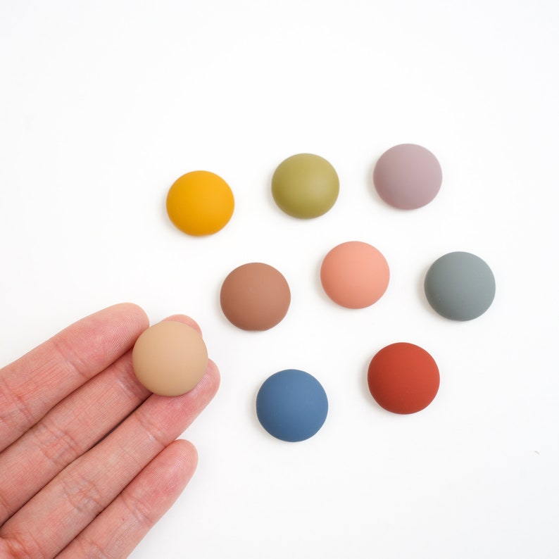 Pu&ograve; includere: Assortimento di nove bottoni rotondi, piatti e colorati in varie tonalit&agrave; di marrone, blu, verde, rosa e giallo. I bottoni sono disposti in cerchio su una superficie bianca.