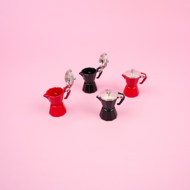 May include: Four miniature espresso makers in red and black with silver accents are arranged on a pink background. Two of the espresso makers have their lids open. The espresso makers have a classic design.
