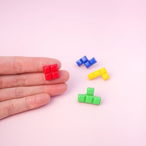 May include: Four colorful Tetris-shaped blocks on a pink background. The blocks are red, blue, yellow, and green. The red block is being held in a hand.
