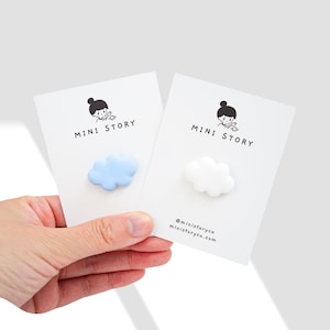 May include: Two white cards with black text and illustrations of a girl with a flower in her hair. The text on the cards reads "MINI STORY".  Each card has a small cloud charm attached to it. One cloud is light blue and the other is white.