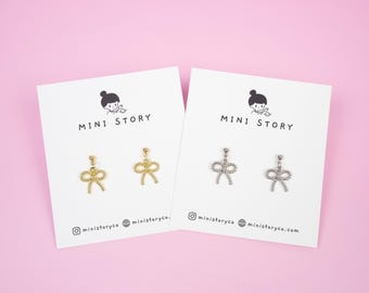Op de afbeelding: Twee paar strikvormige oorbellen, één goudkleurig en één zilverkleurig, gepresenteerd op witte kaarten tegen een roze achtergrond. Elke kaart toont "MINI STORY" en social media details. De oorbellen zijn delicaat en gedetailleerd.