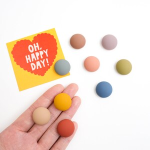 Pu&ograve; includere: Un set di nove bottoni rotondi colorati con un biglietto che dice "Oh, Happy Day!" a forma di cuore. I bottoni sono in tonalit&agrave; di blu, verde, marrone, giallo e rosa.