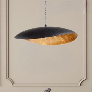 May include: A modern, black and gold pendant light fixture. The top is a textured black, while the underside is a hammered gold. The light hangs from a chain and is suspended in front of a neutral wall. The fixture is approximately 30 inches in length.