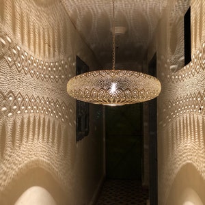 Puede incluir: Una lámpara redonda colgante de color dorado con un patrón decorativo, intrincado y recortado. La lámpara proyecta una sombra con patrón en la pared.