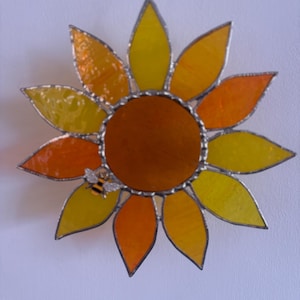 May include: A sunflower-shaped stained glass suncatcher featuring a dark orange circular centre surrounded by alternating yellow and orange petals. A small metal bee figure is attached to one of the petals. The piece is held by a silver chain.