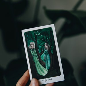 May include: A tarot card with a green and white illustration of two women standing in a forest. The card is titled "The Forest".