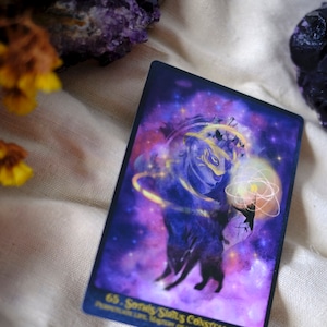May include: A purple and gold tarot card with a starry background. The card features a silhouette of a wolf with a crown and a glowing orb. The text on the card reads "65.  Sandy Stars Constellation".