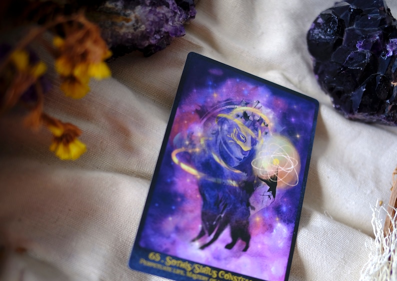 May include: A purple and gold tarot card with a starry background. The card features a silhouette of a wolf with a crown and a glowing orb. The text on the card reads "65.  Sandy Stars Constellation".