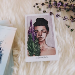 May include: A tarot card with an illustration of a person with their eyes closed, holding a bouquet of purple and green flowers. The card is titled "Sacred Herbs".