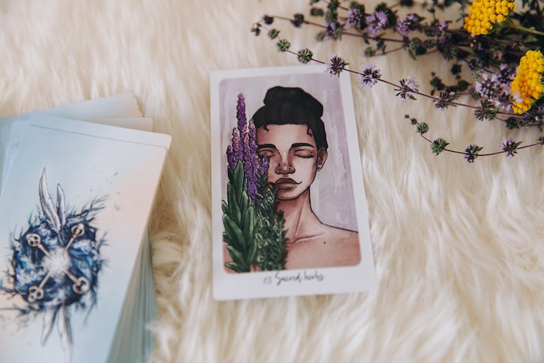 May include: A tarot card with an illustration of a person with their eyes closed, holding a bouquet of purple and green flowers. The card is titled "Sacred Herbs".