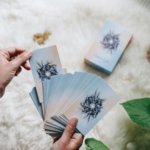 May include: A deck of oracle cards with a blue and white design. The cards are fanned out in a hand, revealing the image of a circle with keys and a blue background. The box for the cards is visible in the background.