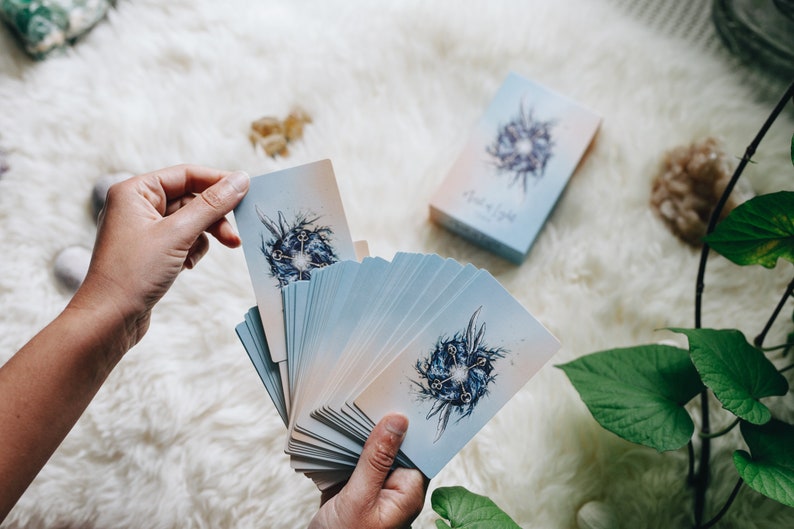 May include: A deck of oracle cards with a blue and white design. The cards are fanned out in a hand, revealing the image of a circle with keys and a blue background. The box for the cards is visible in the background.