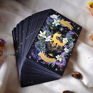 May include: A deck of tarot cards with a black background and gold accents. The top card features a golden unicorn surrounded by white flowers and purple vines. The card also features two golden hands reaching towards the unicorn.