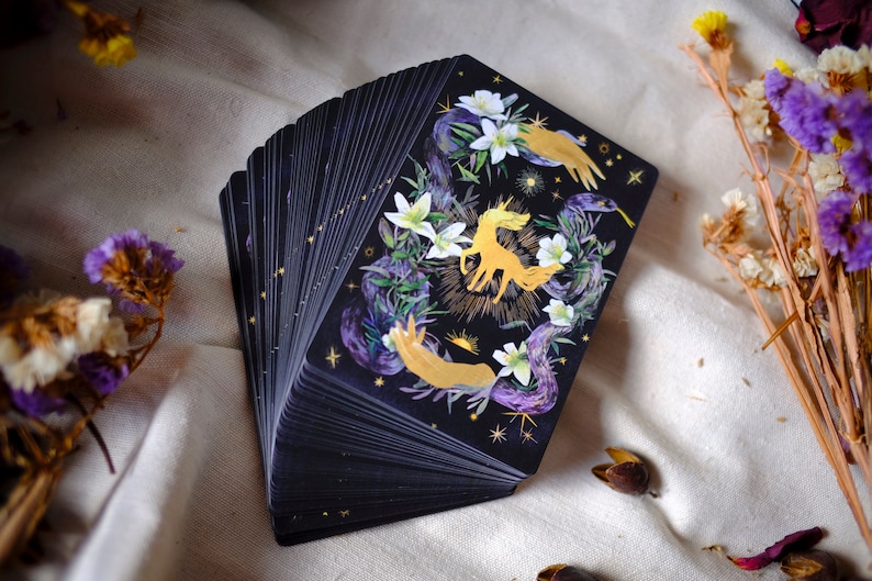 May include: A deck of tarot cards with a black background and gold accents. The top card features a golden unicorn surrounded by white flowers and purple vines. The card also features two golden hands reaching towards the unicorn.