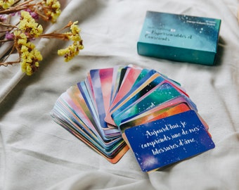May include: A deck of cards with a blue, starry background and the text "Aujourd'hui, je comprends une de mes blessures d'âme."  The cards are fanned out on a white fabric surface.