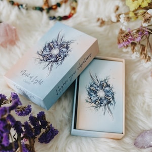 May include: A blue and white box with the text "Nest of Light Oracle" and "Higher your vibrations" on the front. The box is open and the cards inside are visible. The cards have a blue and white watercolor design with a keyhole and keys.