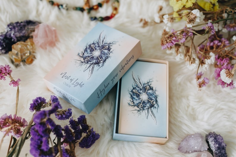 May include: A blue and white box with the text "Nest of Light Oracle" and "Higher your vibrations" on the front. The box is open and the cards inside are visible. The cards have a blue and white watercolor design with a keyhole and keys.