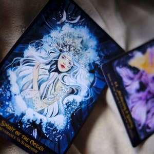 May include: A tarot card with a blue and white illustration of a woman with long white hair wearing a crown. The card is titled "42 Spirit of the Ocean" and the text "Transcendence vs Surrender" is printed below the title.