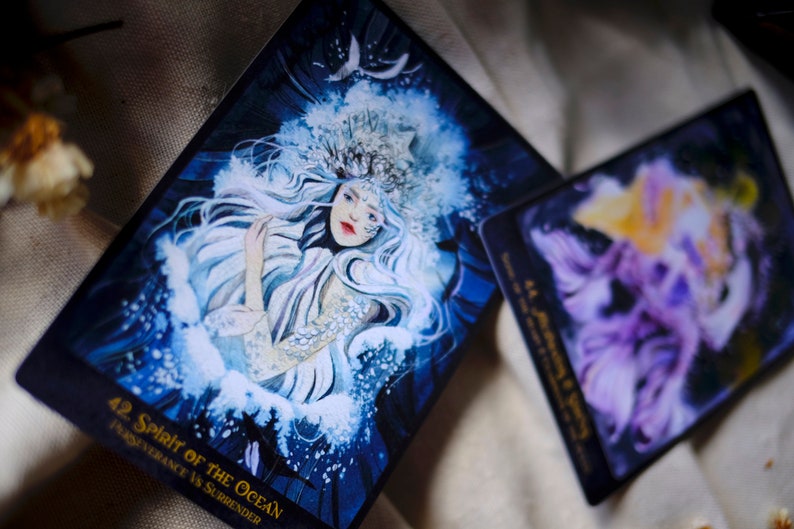 May include: A tarot card with a blue and white illustration of a woman with long white hair wearing a crown. The card is titled "42 Spirit of the Ocean" and the text "Transcendence vs Surrender" is printed below the title.