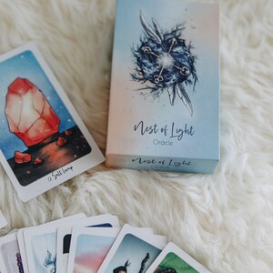 May include: A deck of oracle cards called "Nest of Light Oracle" with a blue and white box. The box features a watercolor illustration of a blue orb with four keys and two wings. The cards are fanned out and show colorful illustrations.