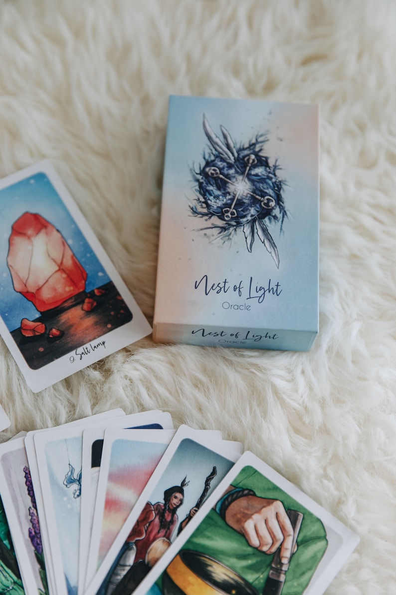 May include: A deck of oracle cards called "Nest of Light Oracle" with a blue and white box. The box features a watercolor illustration of a blue orb with four keys and two wings. The cards are fanned out and show colorful illustrations.