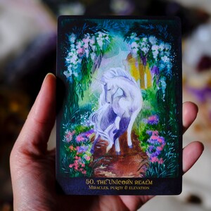 May include: A tarot card with a white unicorn standing on a path in a forest. The card is titled "The Unicorn Realm" and has the text "Miracles, Purity & Elevation" at the bottom.