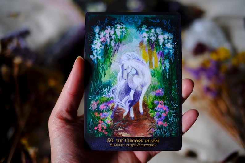 May include: A tarot card with a white unicorn standing on a path in a forest. The card is titled "The Unicorn Realm" and has the text "Miracles, Purity & Elevation" at the bottom.