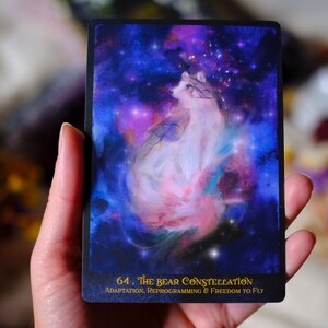 May include: A tarot card with a white wolf-like creature in a swirling nebula of pink, blue, and purple. The card is titled "64. The Bear Constellation" with the text "Adaptation, Reprogramming & Freedom to Fly" below.
