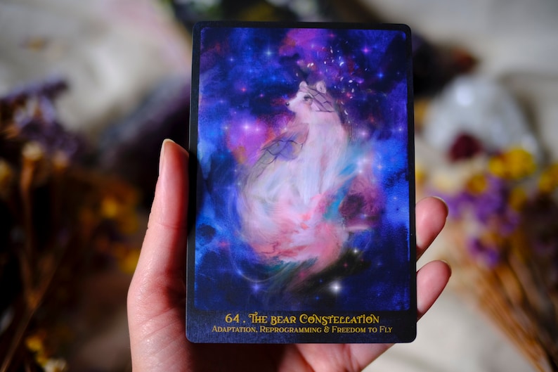 May include: A tarot card with a white wolf-like creature in a swirling nebula of pink, blue, and purple. The card is titled "64. The Bear Constellation" with the text "Adaptation, Reprogramming & Freedom to Fly" below.