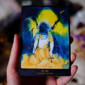 May include: A tarot card with a dark blue background and a yellow and blue watercolor illustration of a figure with wings. The figure is wearing a white robe and a gold belt. The card is titled "21. Ra Power & Creation".