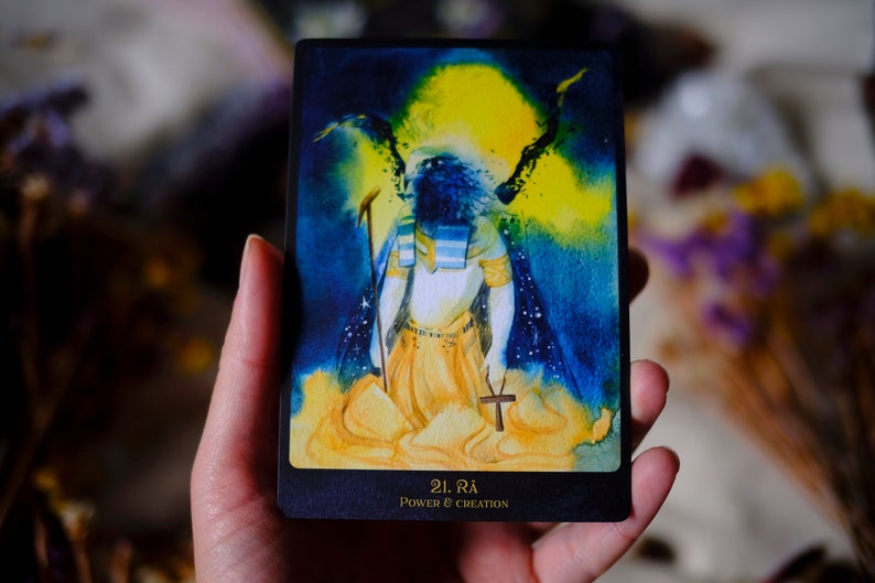 May include: A tarot card with a dark blue background and a yellow and blue watercolor illustration of a figure with wings. The figure is wearing a white robe and a gold belt. The card is titled "21. Ra Power & Creation".