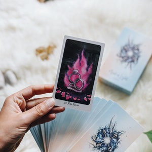 May include: A hand holds a tarot card with a pink flame and two silver rings. The card is labeled "Rose Flame". The card is part of a deck of tarot cards with blue and white backgrounds.