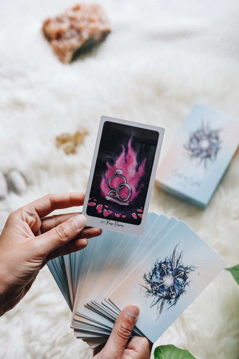 May include: A hand holds a tarot card with a pink flame and two silver rings. The card is labeled "Rose Flame". The card is part of a deck of tarot cards with blue and white backgrounds.
