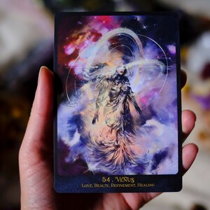May include: A tarot card with a purple, pink, and blue background. A woman with long hair is standing in the center of the card. The card is labeled "54. Venus" and "LOVE, BEAUTY, REFINEMENT, HEALING".
