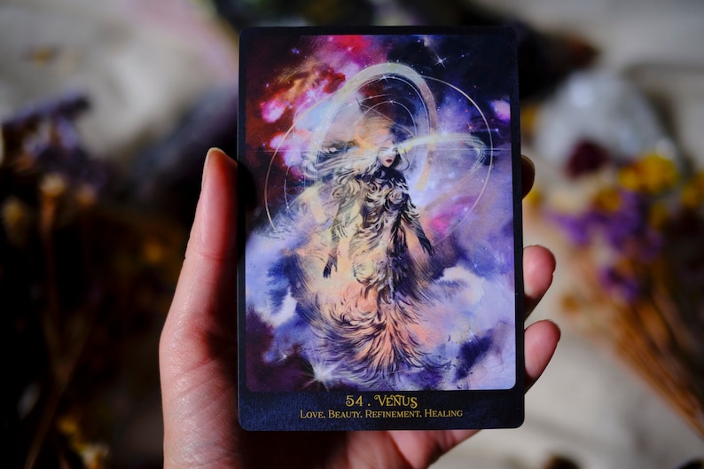 May include: A tarot card with a purple, pink, and blue background. A woman with long hair is standing in the center of the card. The card is labeled "54. Venus" and "LOVE, BEAUTY, REFINEMENT, HEALING".