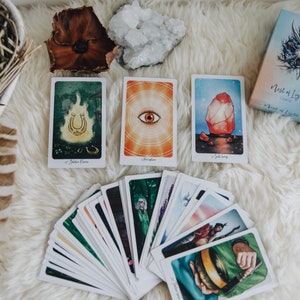 May include: A deck of tarot cards with a white background. The cards are fanned out and show colorful illustrations. The box for the deck is in the upper right corner and says "Nest of Light Oracle".