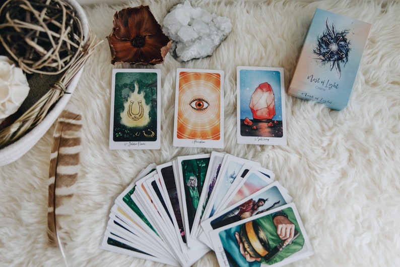 May include: A deck of tarot cards with a white background. The cards are fanned out and show colorful illustrations. The box for the deck is in the upper right corner and says "Nest of Light Oracle".