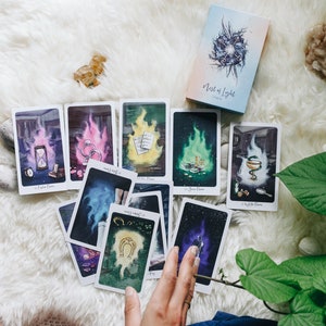 May include: A spread of tarot cards on a white fuzzy surface with a green plant in the background. The cards are from the "Nest of Light" deck and feature colorful illustrations.