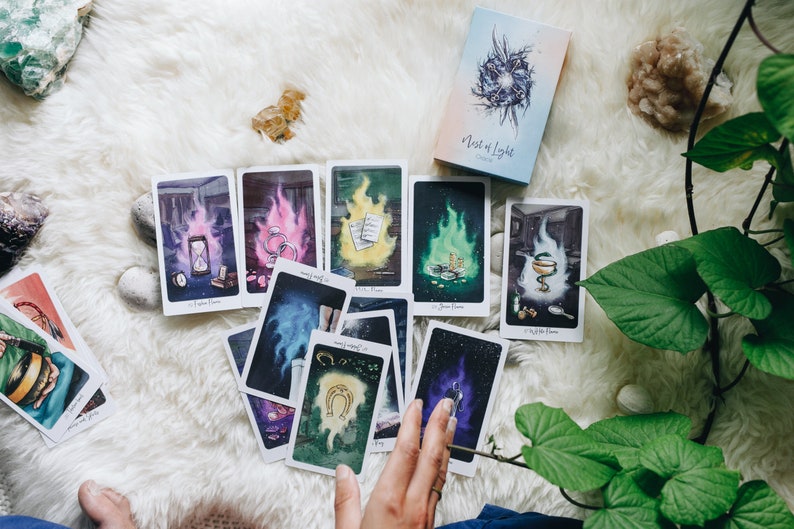 May include: A spread of tarot cards on a white fuzzy surface with a green plant in the background. The cards are from the "Nest of Light" deck and feature colorful illustrations.