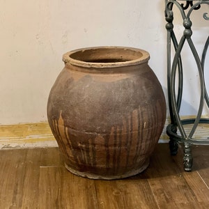 May include: A large, brown, earthenware pot with a wide mouth and a slightly rounded body. The pot has a rustic, aged appearance with streaks of darker brown glaze.