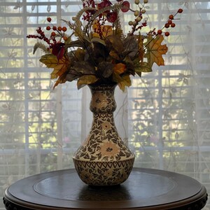 May include: A white ceramic vase with a floral pattern and a heart border, filled with a fall-themed arrangement of red berries, brown leaves, and yellow flowers. The vase is sitting on a dark brown wooden table.