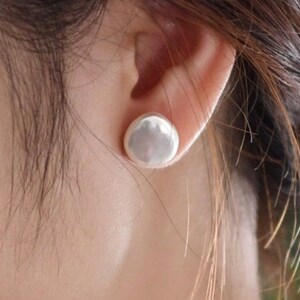 May include: A close-up of a silver-colored, round stud earring. The earring is smooth and reflective, with a slightly domed shape. The earring is set against the backdrop of a person's ear and hair.