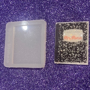 May include: A clear silicone mould shaped like a composition notebook. The mould is next to a finished resin piece that looks like a composition notebook with the words "Composition" and "Mrs. Morin" on the front.