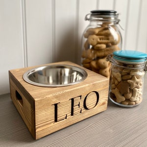 May include: A wooden dog bowl stand with a stainless steel bowl and the name "LEO" engraved on the front. The stand is made of natural wood and has a rustic look. Two glass jars filled with dog treats are in the background.